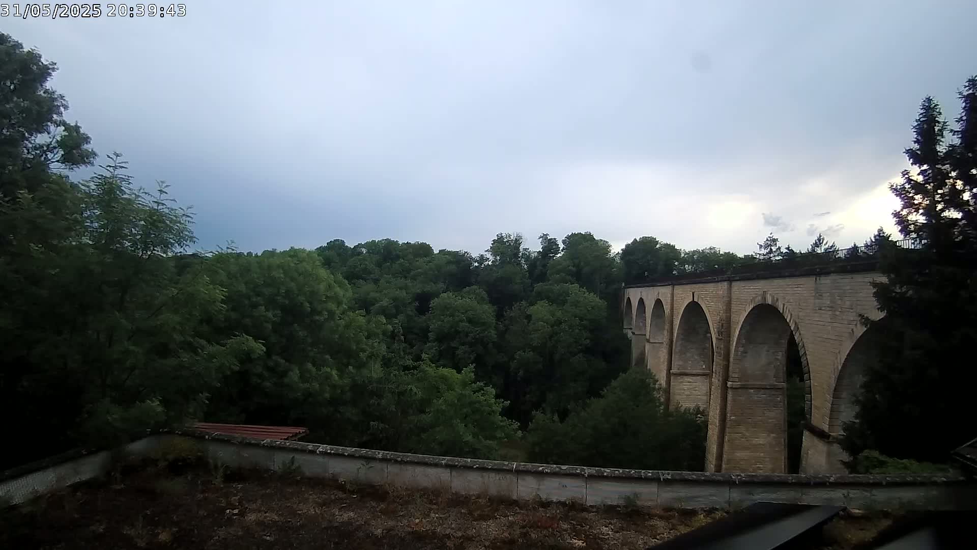 A stone arched bridge stretches across a valley filled with lush green trees under a cloudy sky.