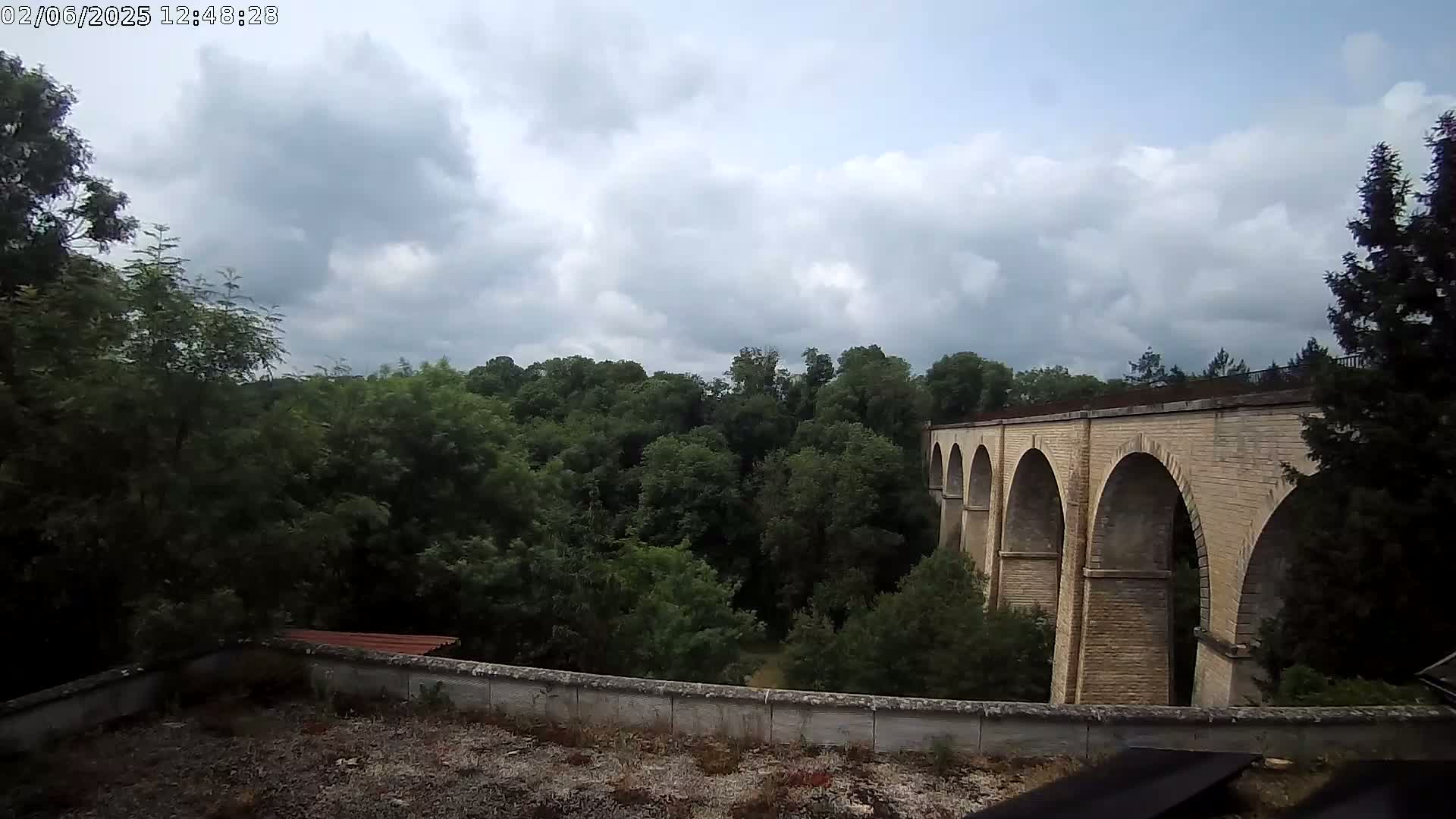 A stone viaduct with multiple arches is visible beyond a line of lush green trees under a partly cloudy sky.