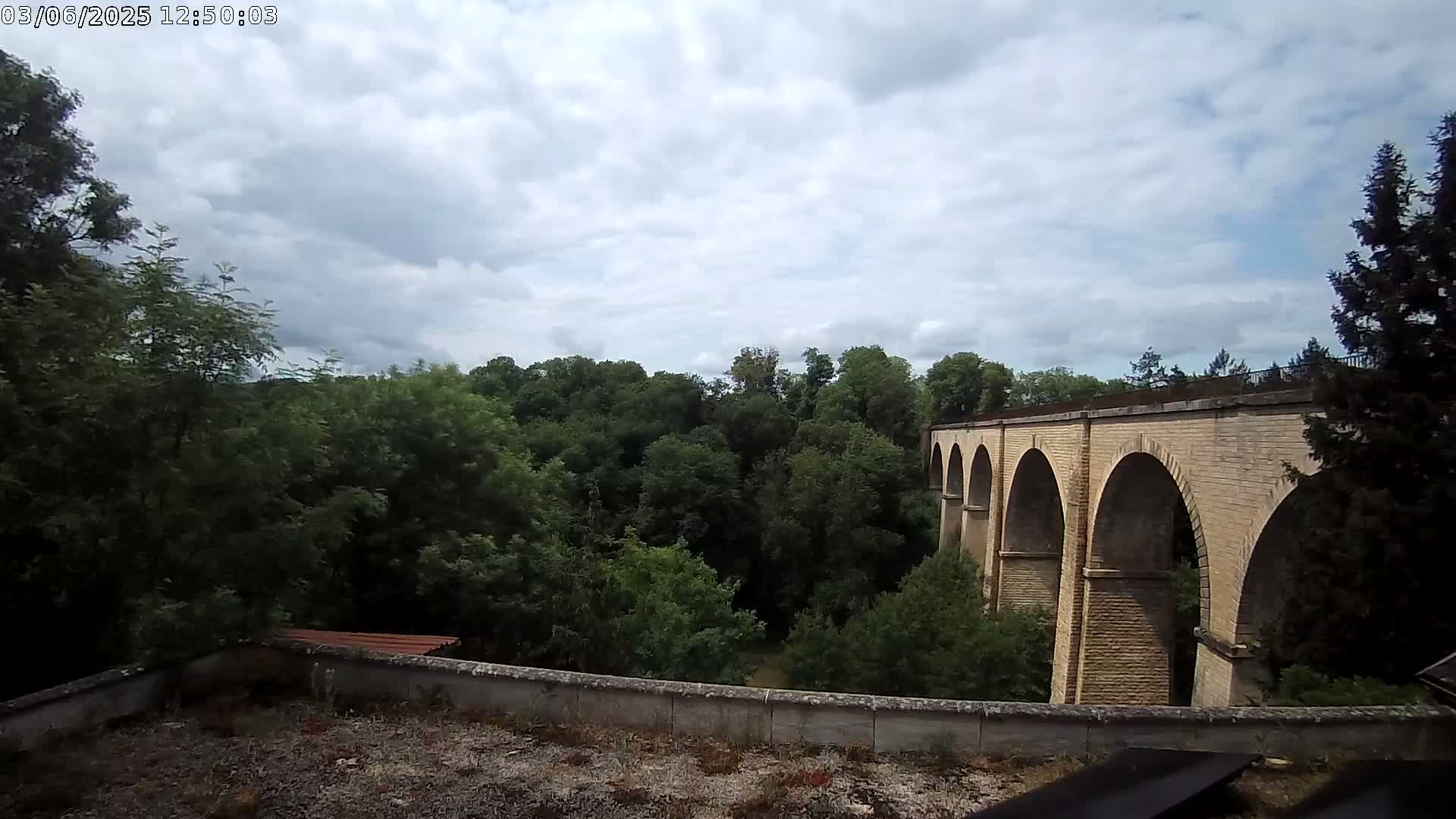 A stone viaduct with multiple arches is seen over a lush green valley under a partly cloudy sky.