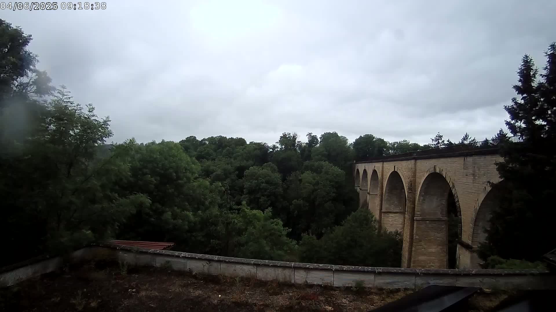 A stone arched bridge is partially visible behind a wall and lush green trees under an overcast sky.