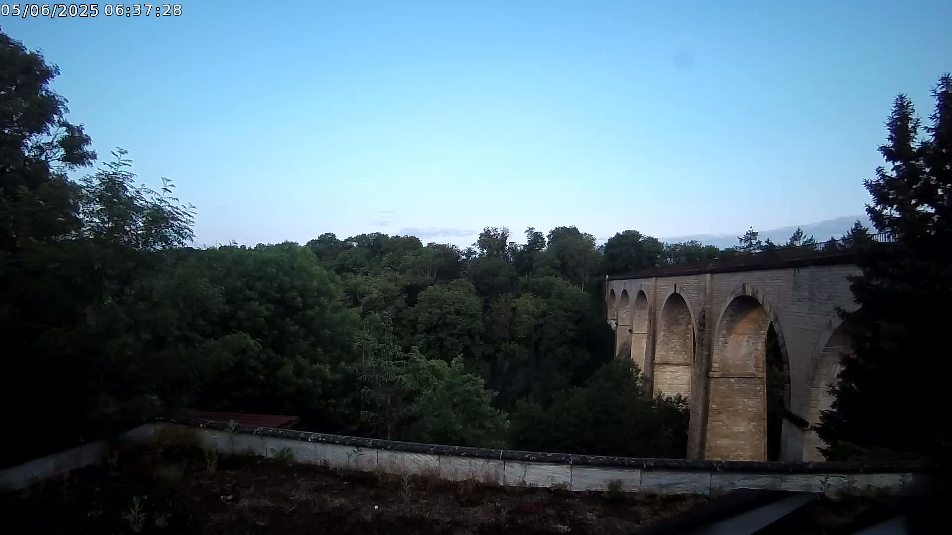 A stone viaduct with multiple arches is visible beyond a line of leafy green trees under a clear blue sky.