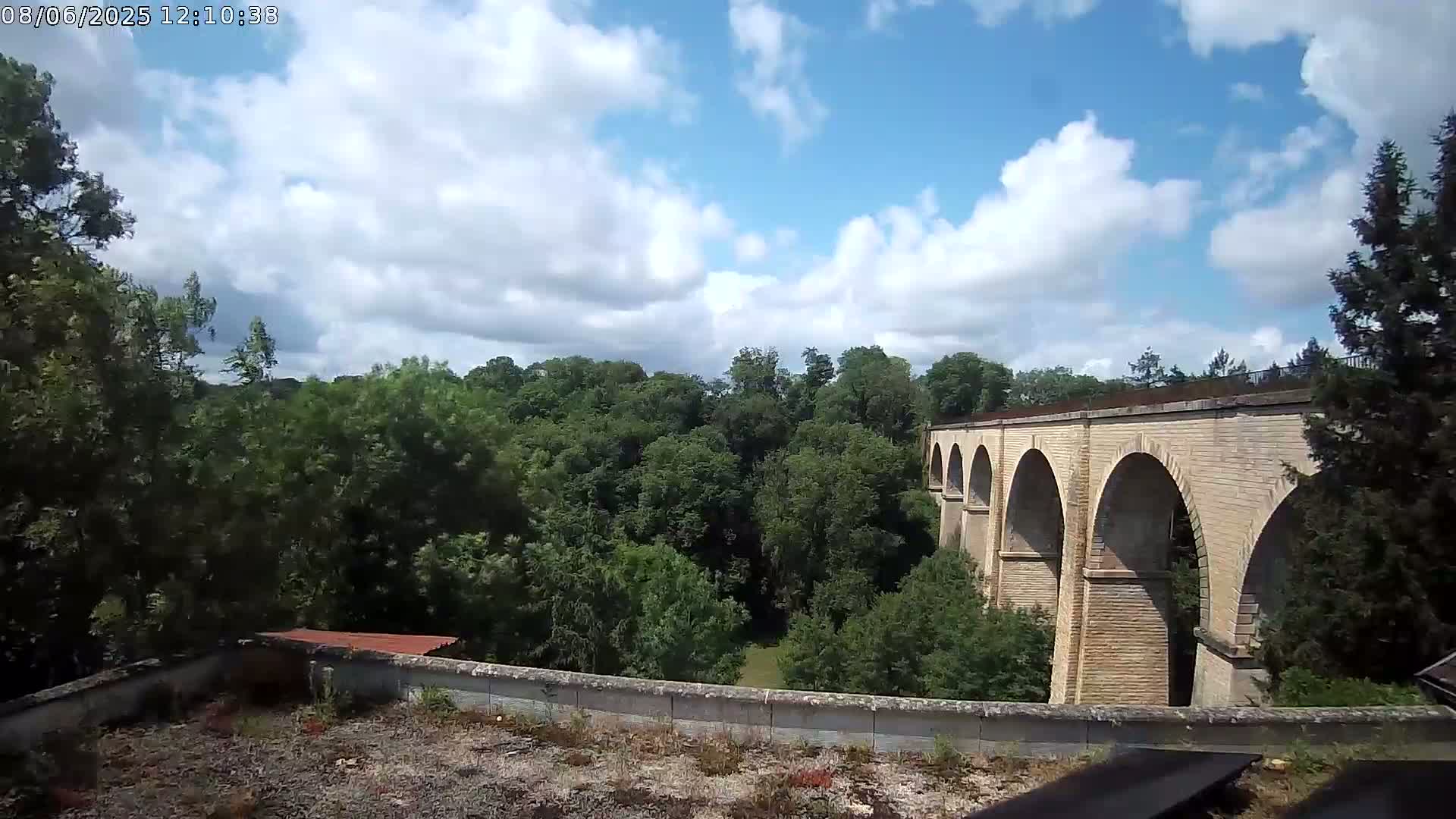 A stone viaduct with multiple arches stretches across a verdant valley under a partly cloudy sky.