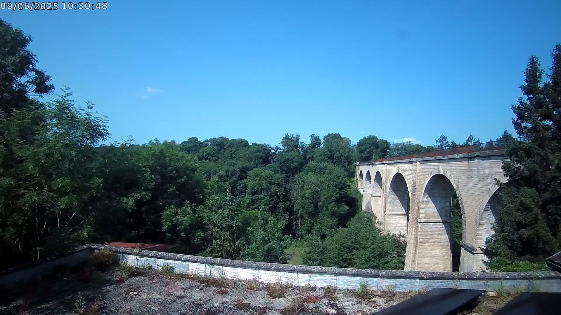 A stone viaduct with multiple arches stretches across a valley of lush green trees under a clear blue sky.