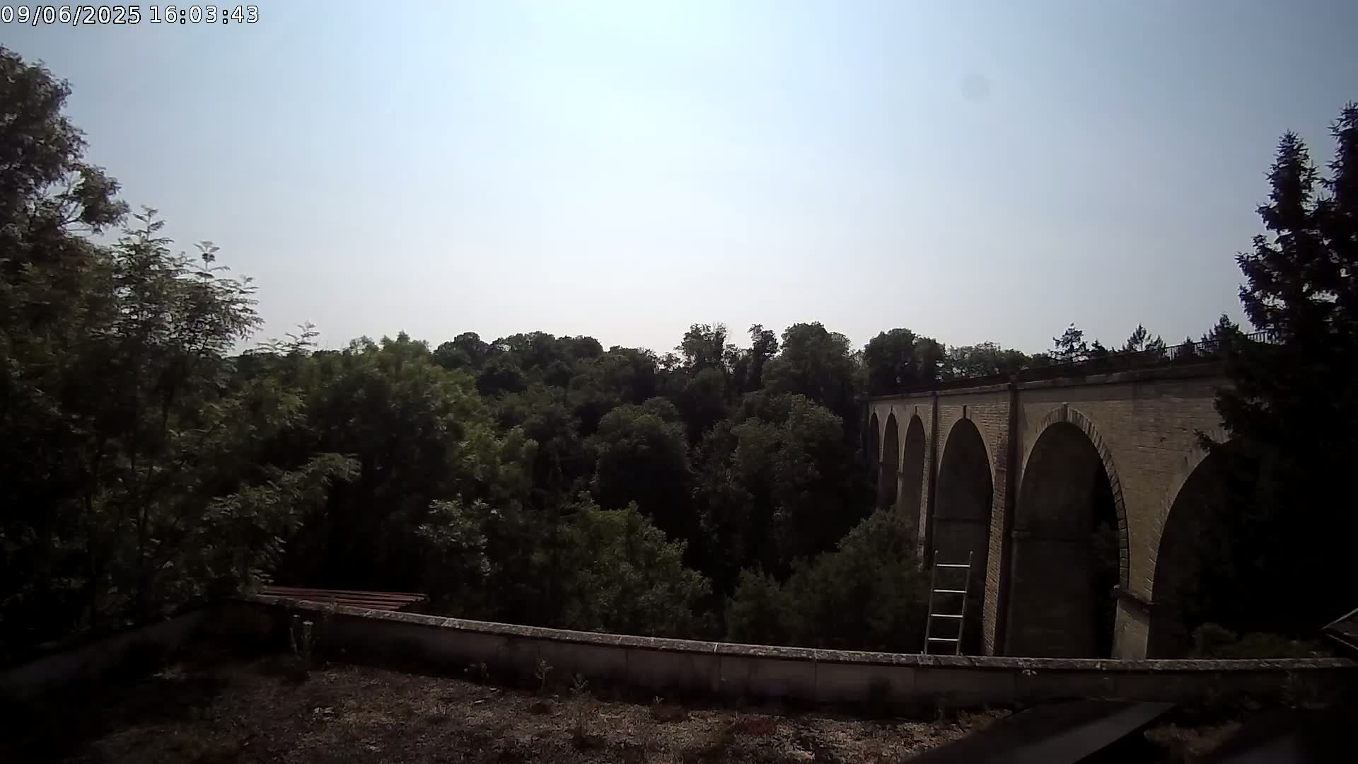 A stone viaduct with multiple arches is seen beyond a line of leafy green trees under a clear, bright sky.