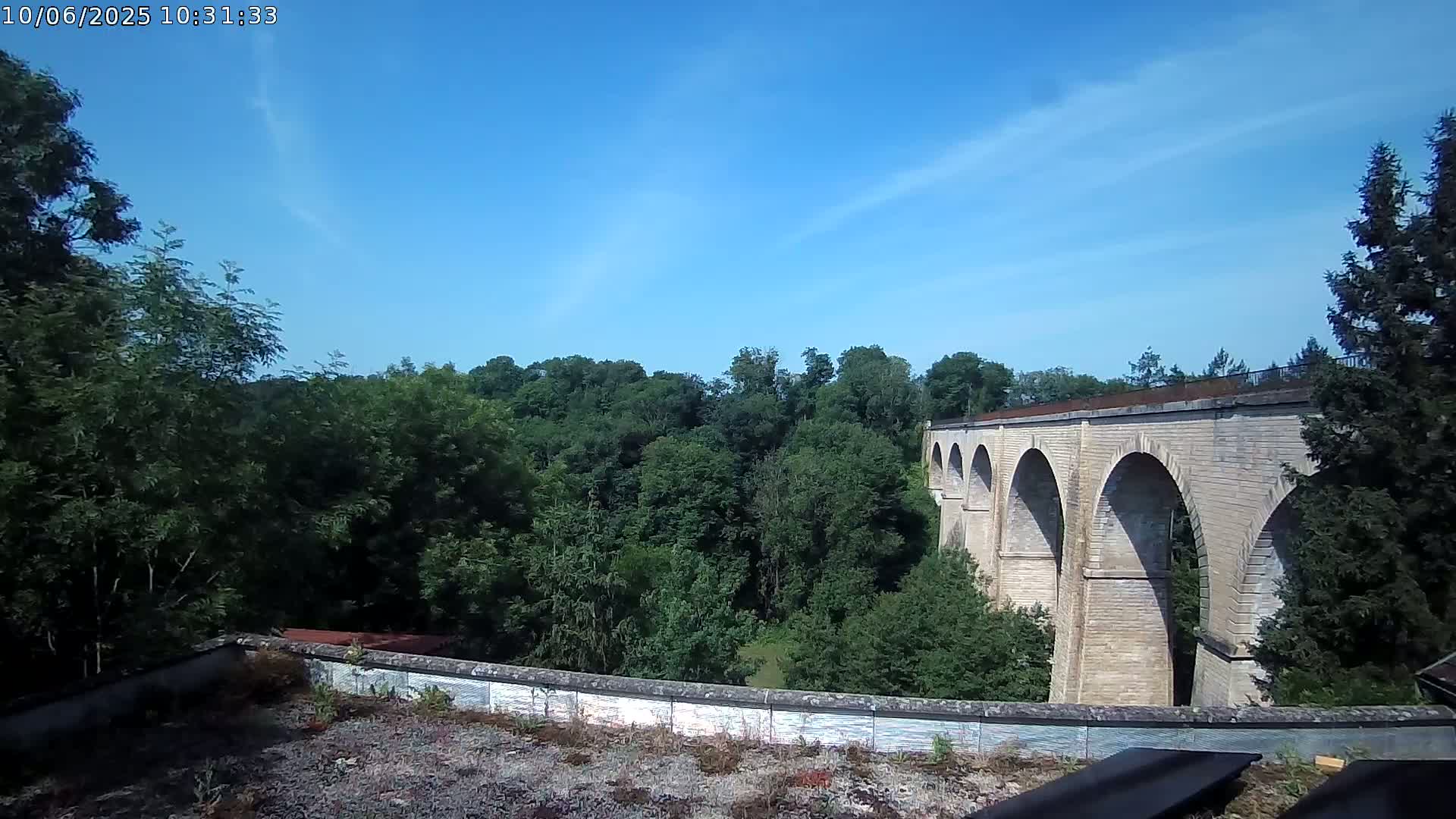 A stone viaduct stretches across a valley of lush green trees under a mostly clear blue sky.