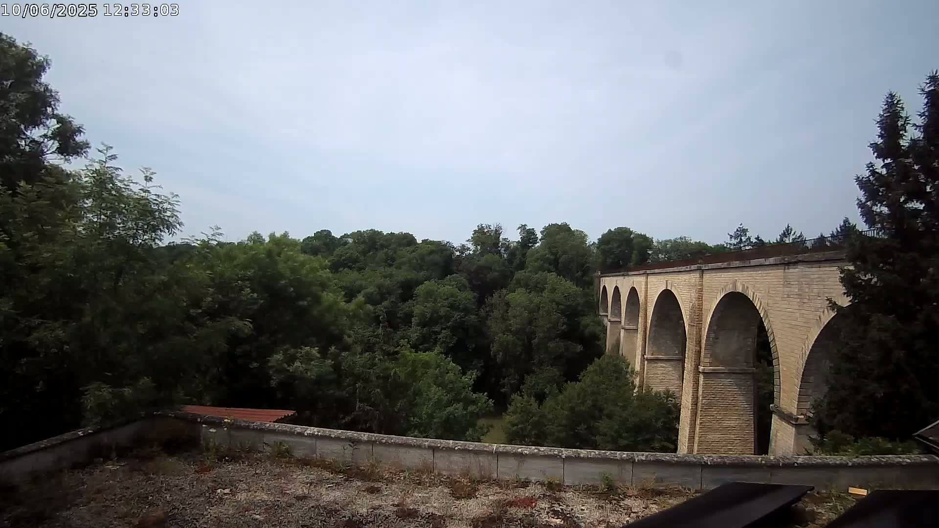 A stone viaduct with multiple arches is visible beyond a line of lush green trees under a cloudy sky.