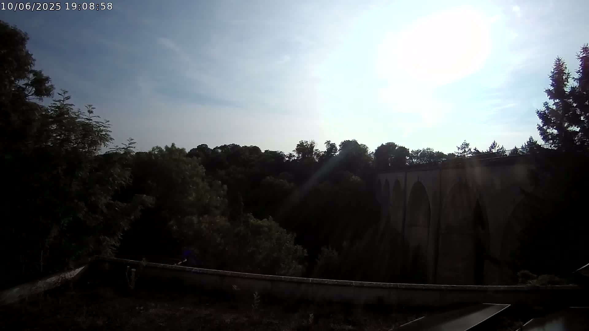 A stone bridge with arched openings is partially visible beyond a line of dark green trees under a mostly sunny sky.