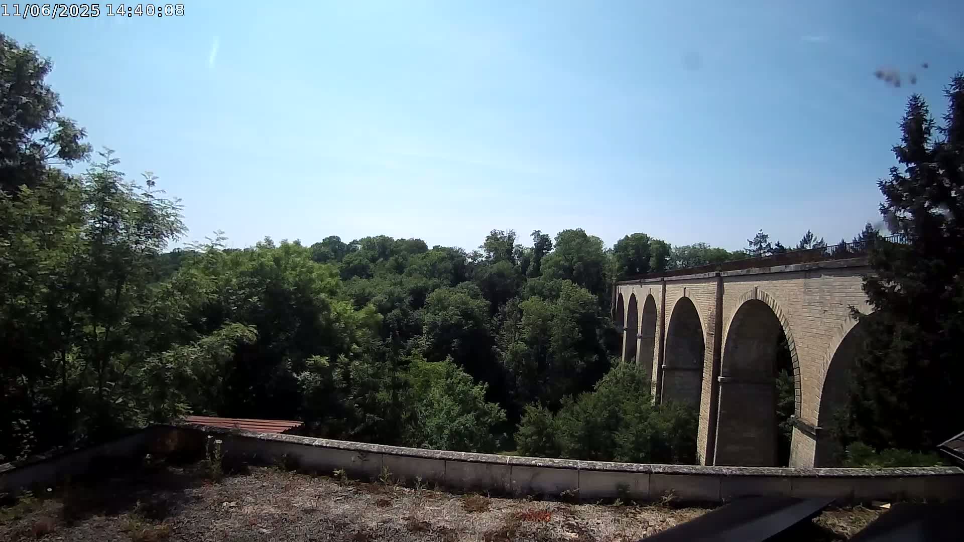 A stone viaduct with multiple arches is visible beyond a line of leafy green trees under a clear blue sky.