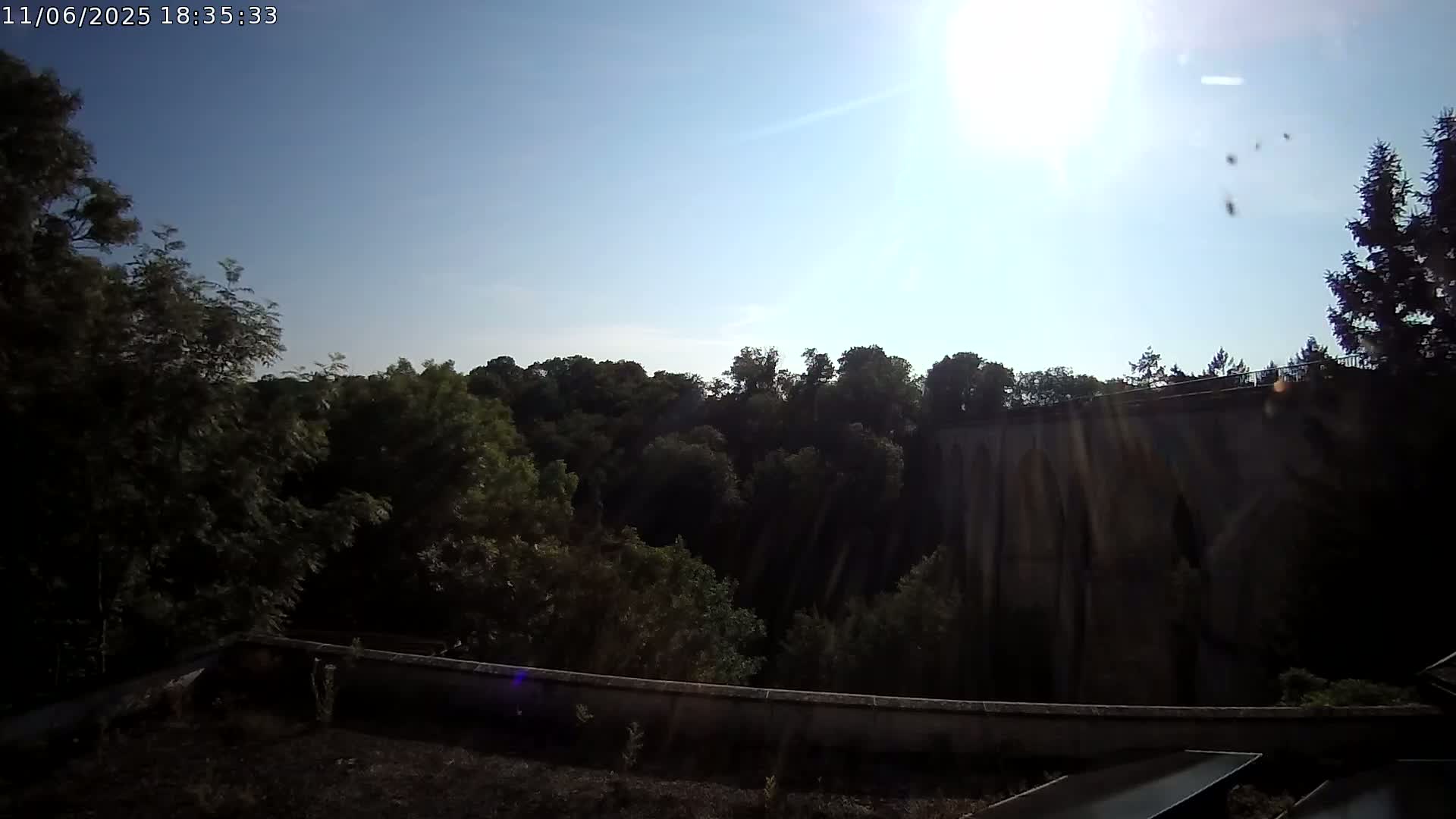 A sunny day reveals a view of lush green trees and a large stone structure, possibly a bridge or aqueduct, from a slightly elevated vantage point.