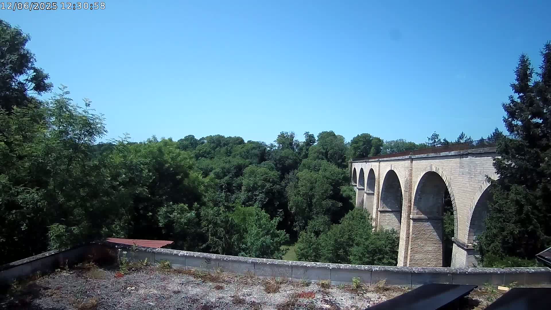 A stone viaduct with multiple arches is visible beyond a line of lush green trees under a clear blue sky.