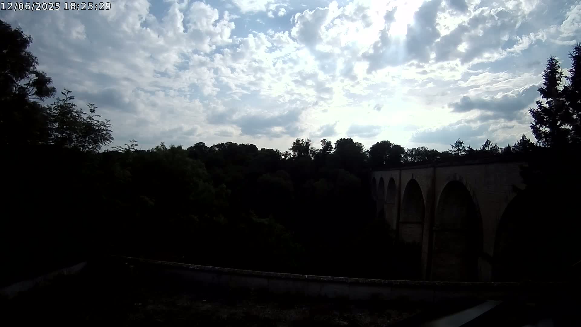 A stone arched bridge is partially visible behind a dark line of trees under a partly sunny sky with fluffy clouds.