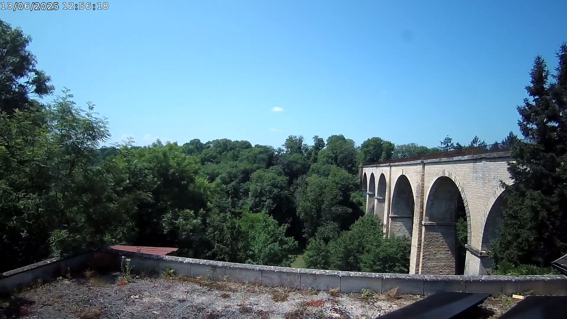 A stone viaduct with multiple arches is visible beyond a line of green trees under a clear blue sky.