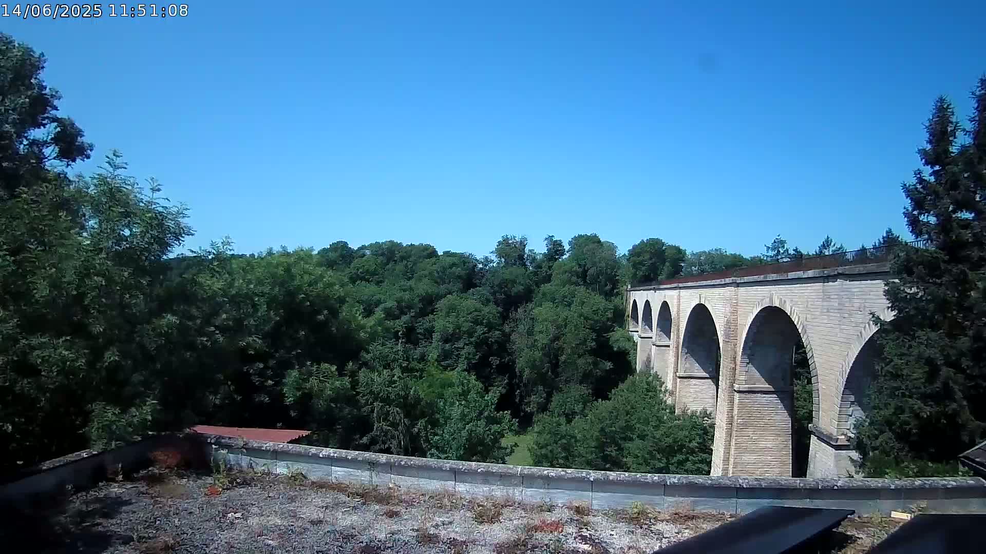 A stone viaduct with multiple arches is visible beyond a rooftop and a lush green forest under a clear blue sky.