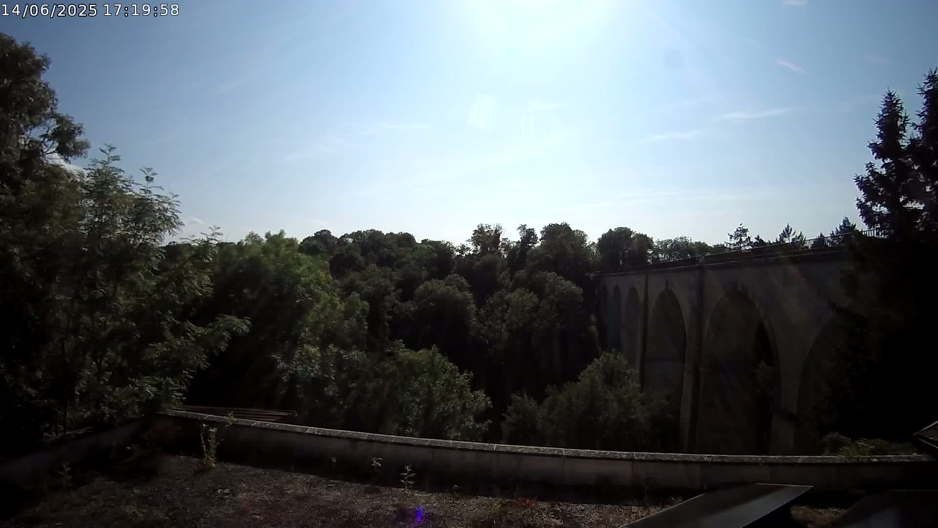 A stone arched bridge is partially visible beyond a wall and a line of leafy green trees under a mostly sunny sky.