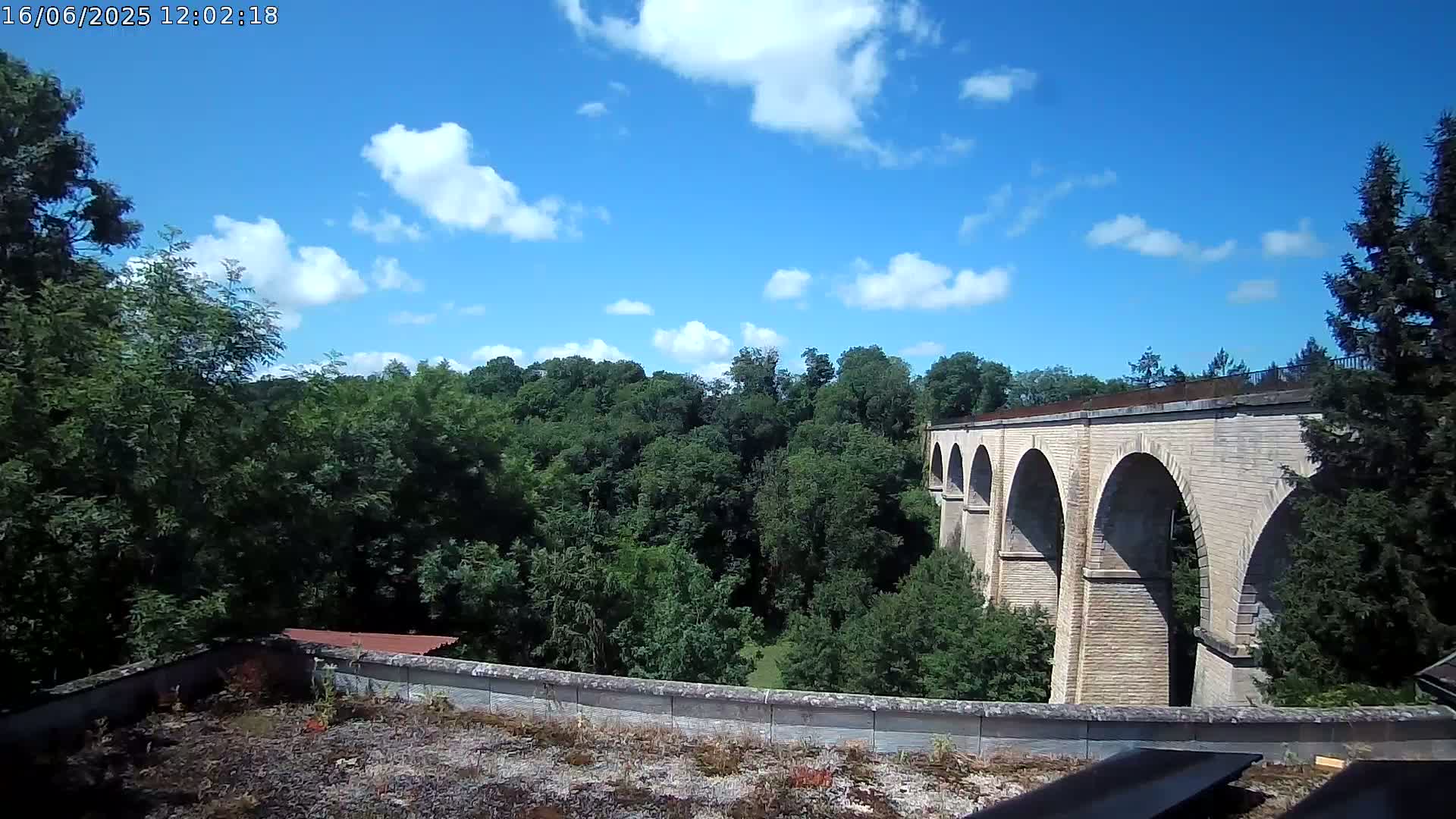 A stone viaduct with multiple arches stretches across a lush green valley under a mostly sunny sky with scattered clouds.