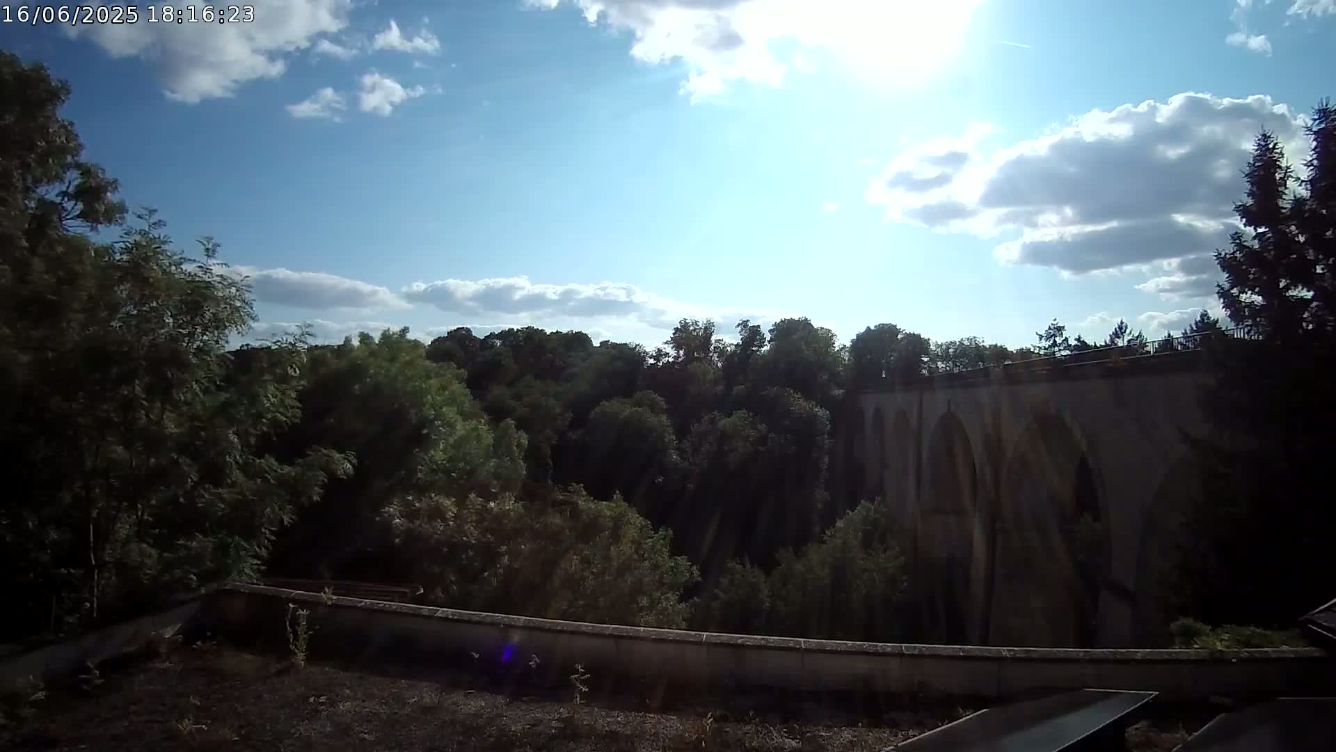 A stone bridge with arched openings is visible beyond a line of leafy green trees under a partly sunny sky.
