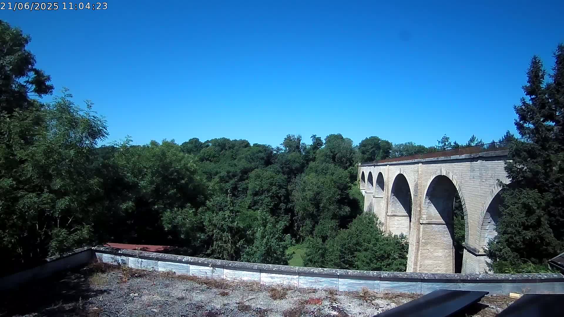 A stone viaduct with multiple arches stretches across a lush green valley under a clear blue sky.