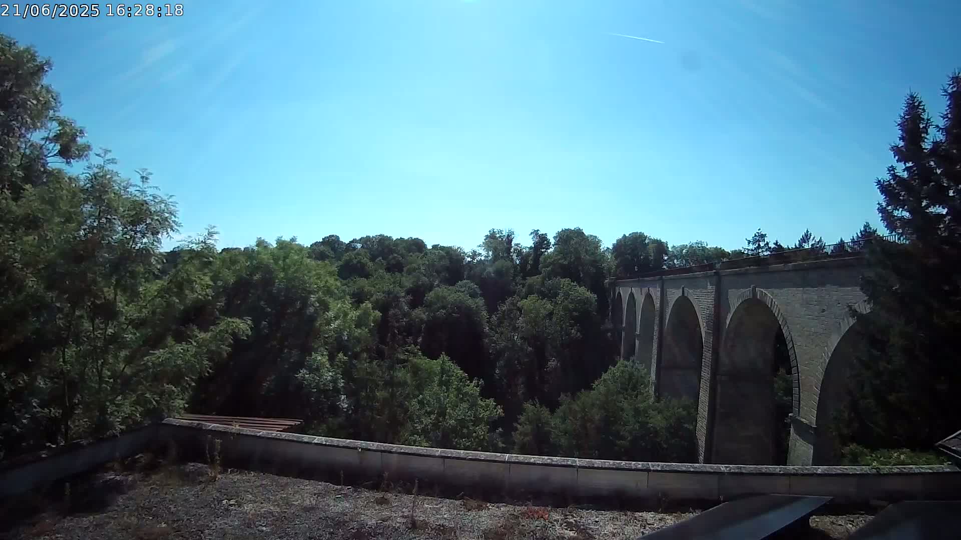 A stone arched bridge is visible beyond a lush green forest under a clear blue sky.
