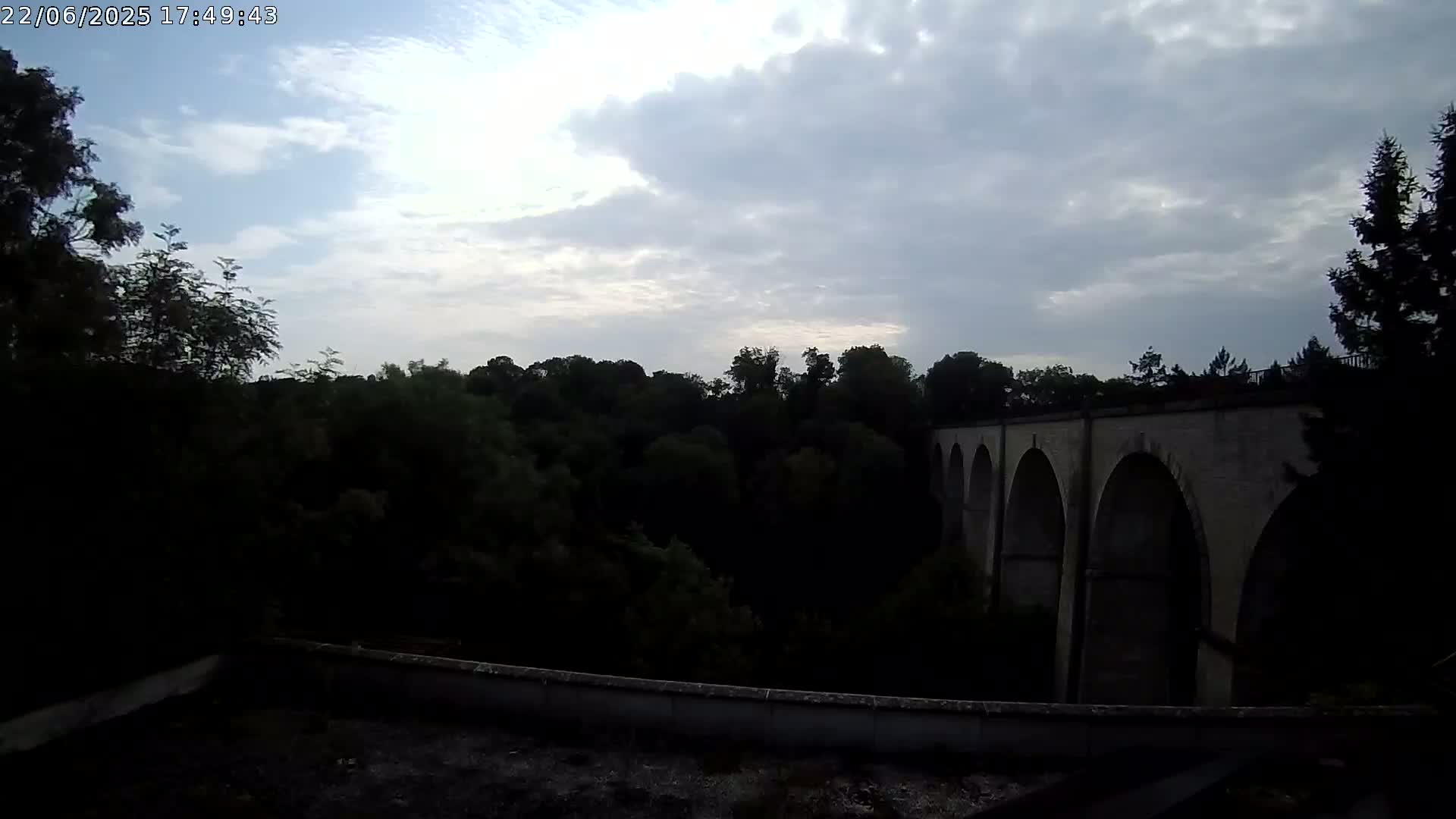 A stone viaduct with multiple arches is visible beyond a dark line of trees under a partly cloudy sky.