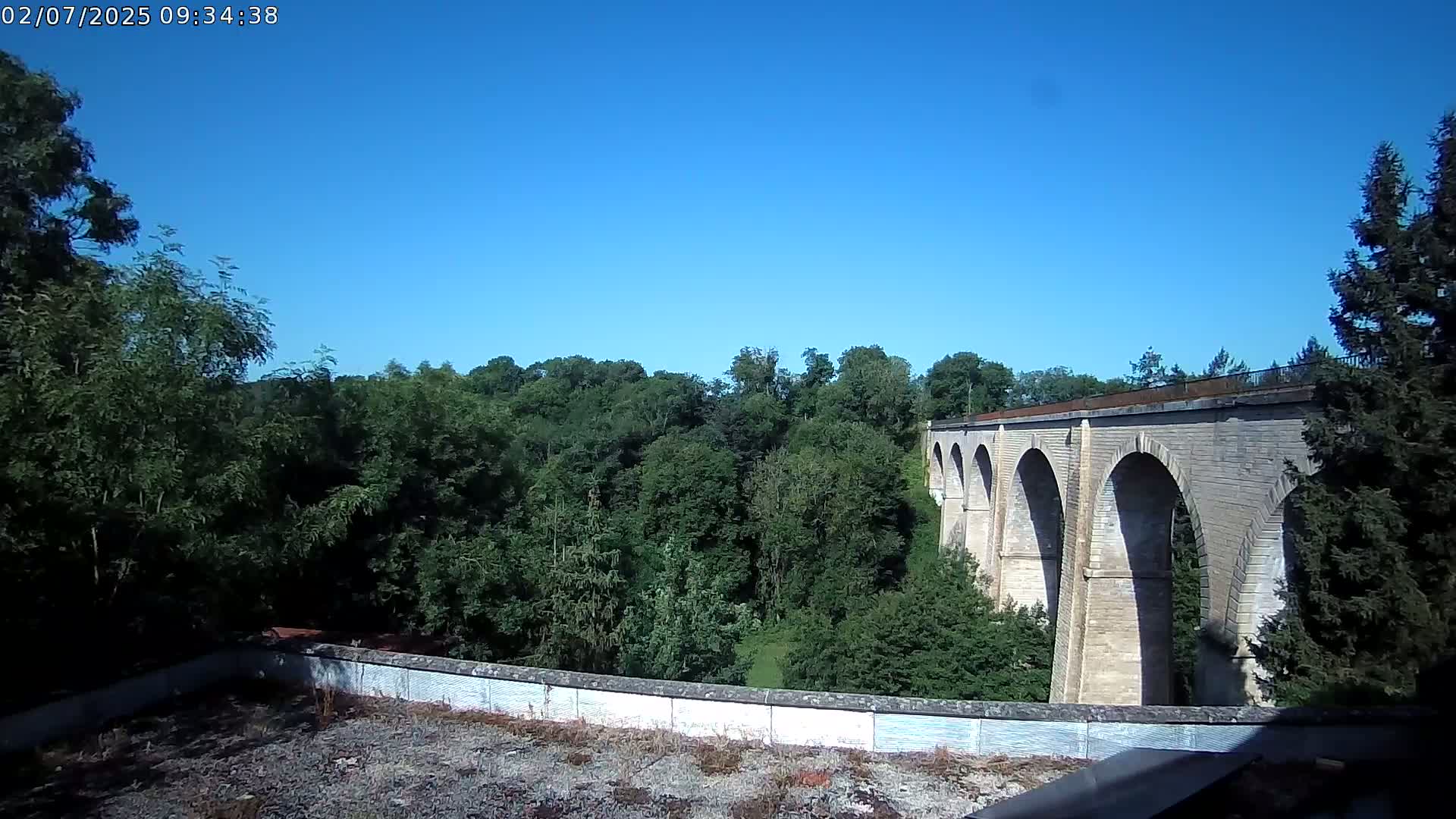 A stone viaduct with multiple arches stretches across a lush green valley under a clear blue sky.