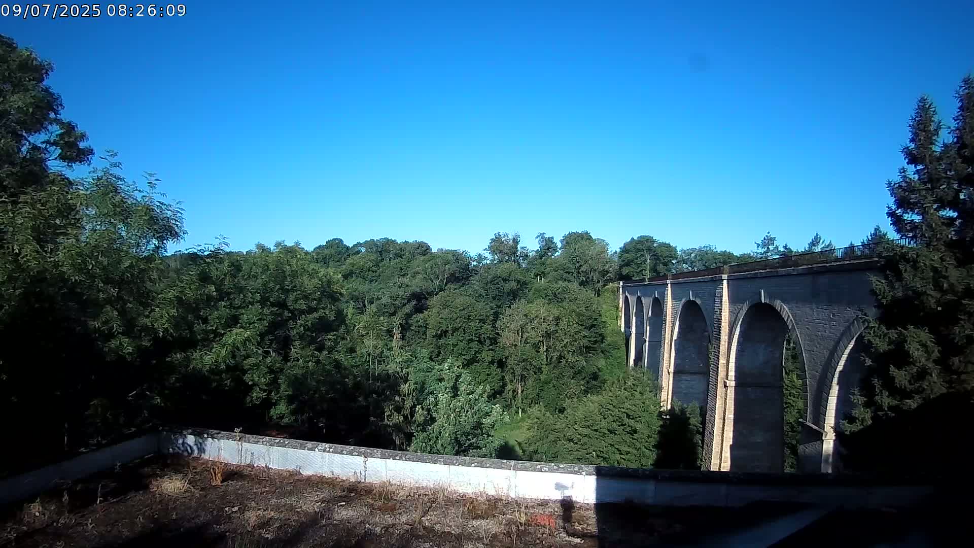 A stone viaduct with multiple arches stretches across a lush green valley under a clear blue sky.