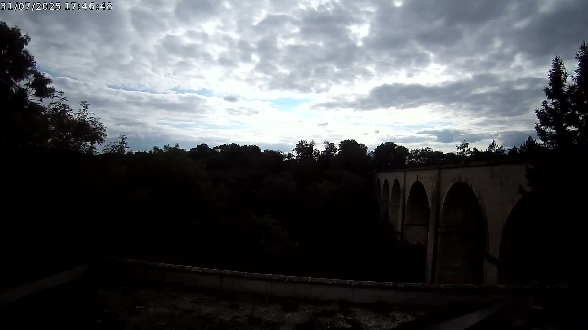 A stone arched bridge is visible through a mostly cloudy sky, partially obscured by dark trees.