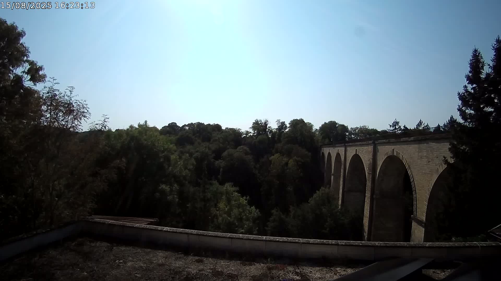 A stone arched bridge is visible through a mostly cloudy sky, partially obscured by dark trees.