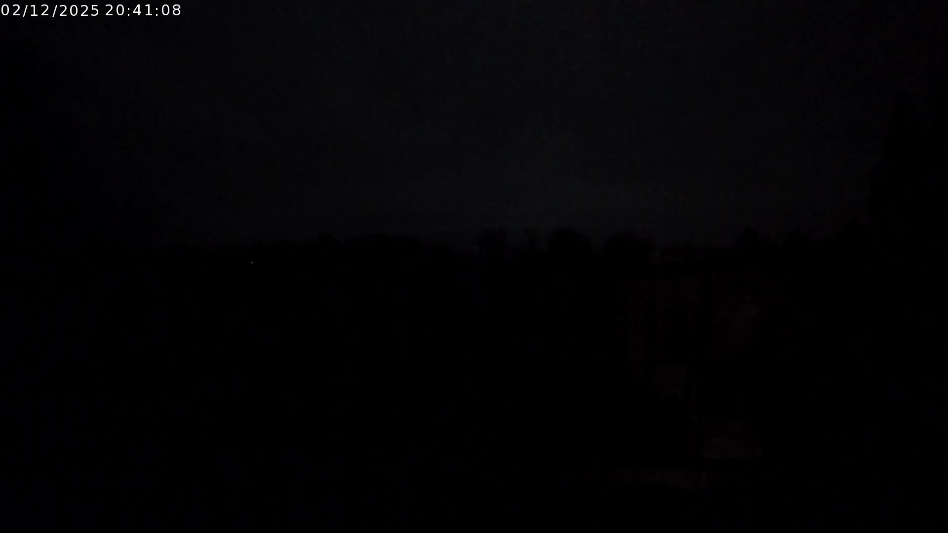 The image depicts an extremely dark outdoor scene on an overcast night, with a faint horizon line of distant trees or structures and a tiny bright dot barely discernible against the dark sky.
