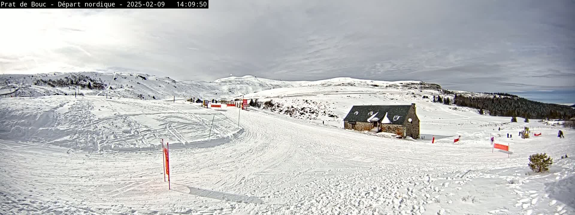 A snow-covered landscape with a small stone building and several people skiing or snowboarding under a cloudy sky.