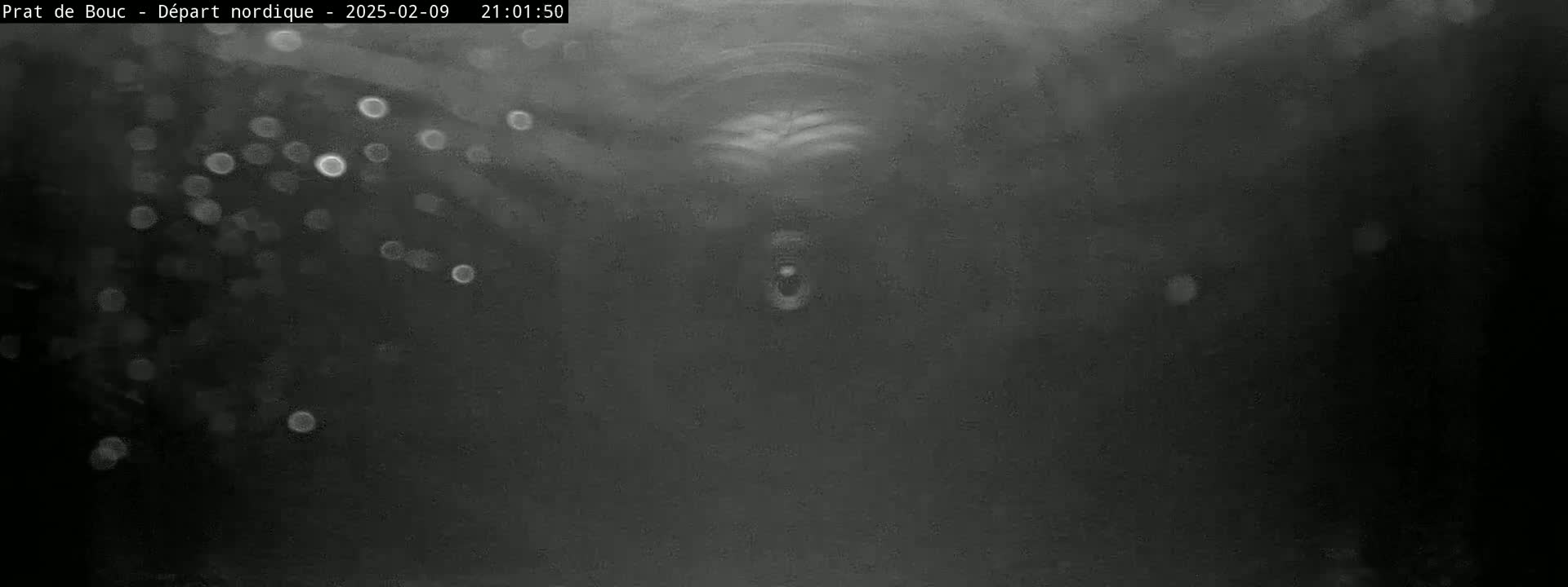 A blurry, dark grayscale image shows a dark background with light, out-of-focus spots possibly depicting falling snow or rain at night.