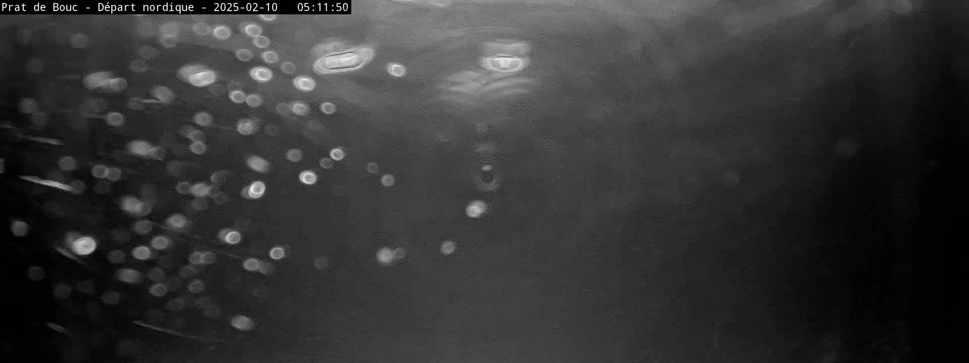 The image shows a dark, blurry scene with numerous out-of-focus white spots appearing to fall from the top, suggesting it is snowing at night.