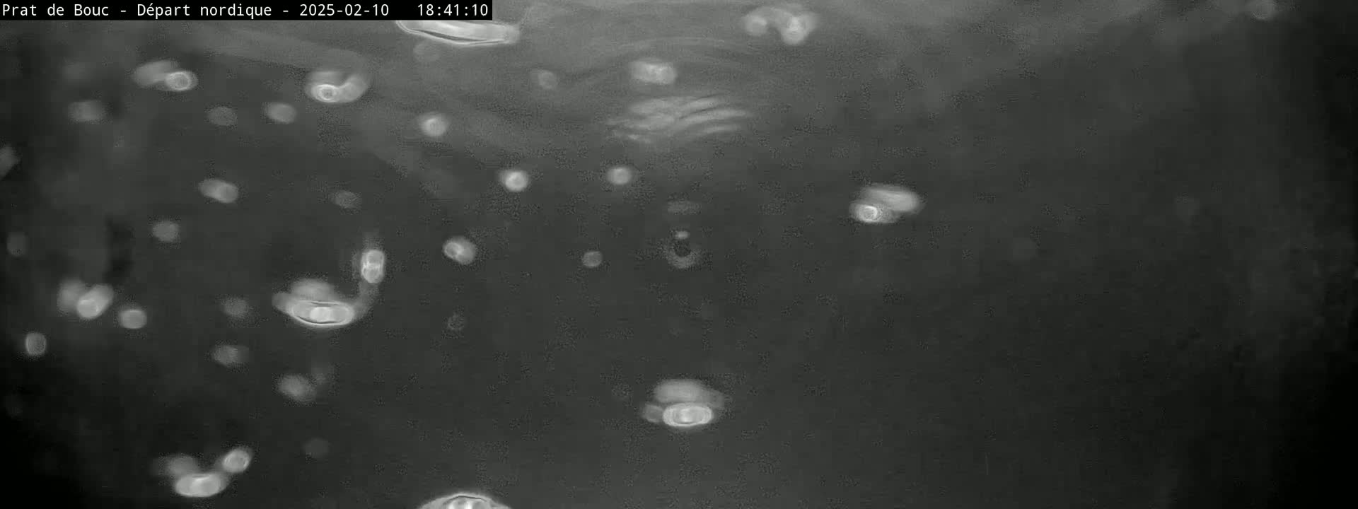 A dark, blurry image shows numerous out-of-focus light-colored spots scattered against a dark gray background, possibly depicting raindrops on a window at night.