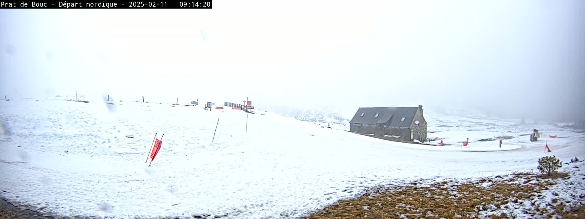 A snowy landscape featuring a long, low stone building, a winding road, and several small red flags in the foggy weather.