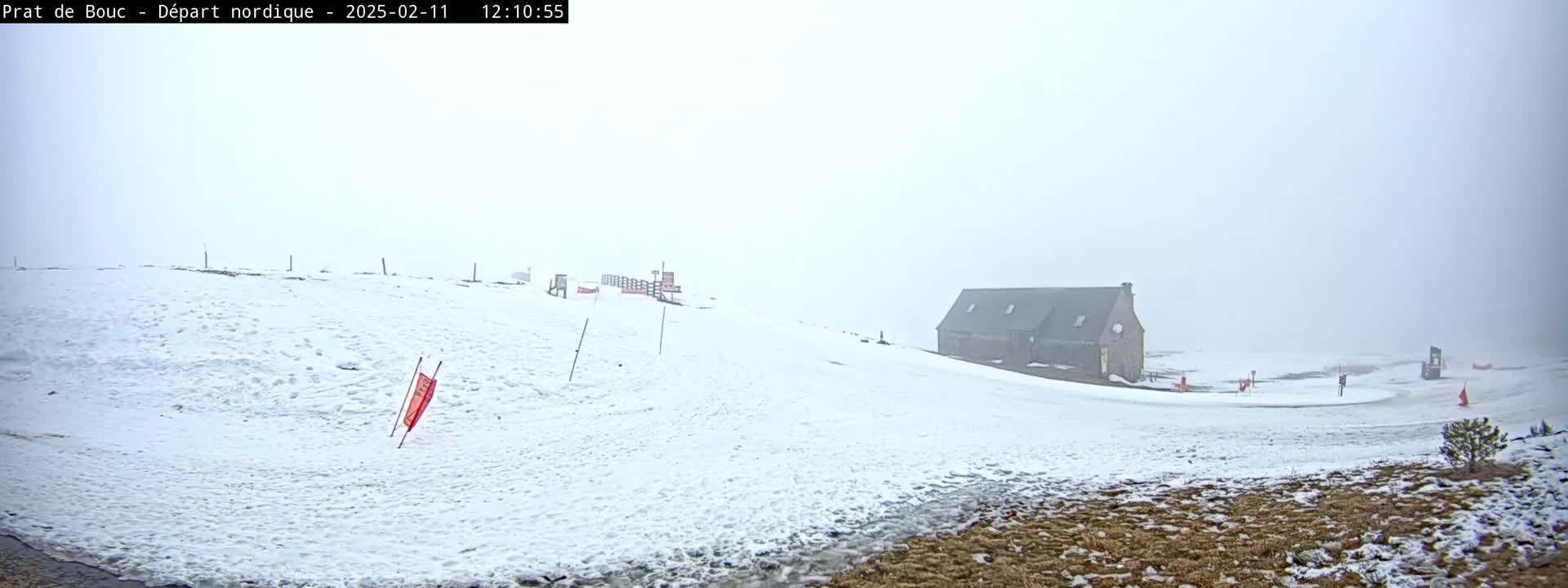 A snow-covered landscape, shrouded in fog, features a small building on a hillside with sparse vegetation visible in the lower right corner.
