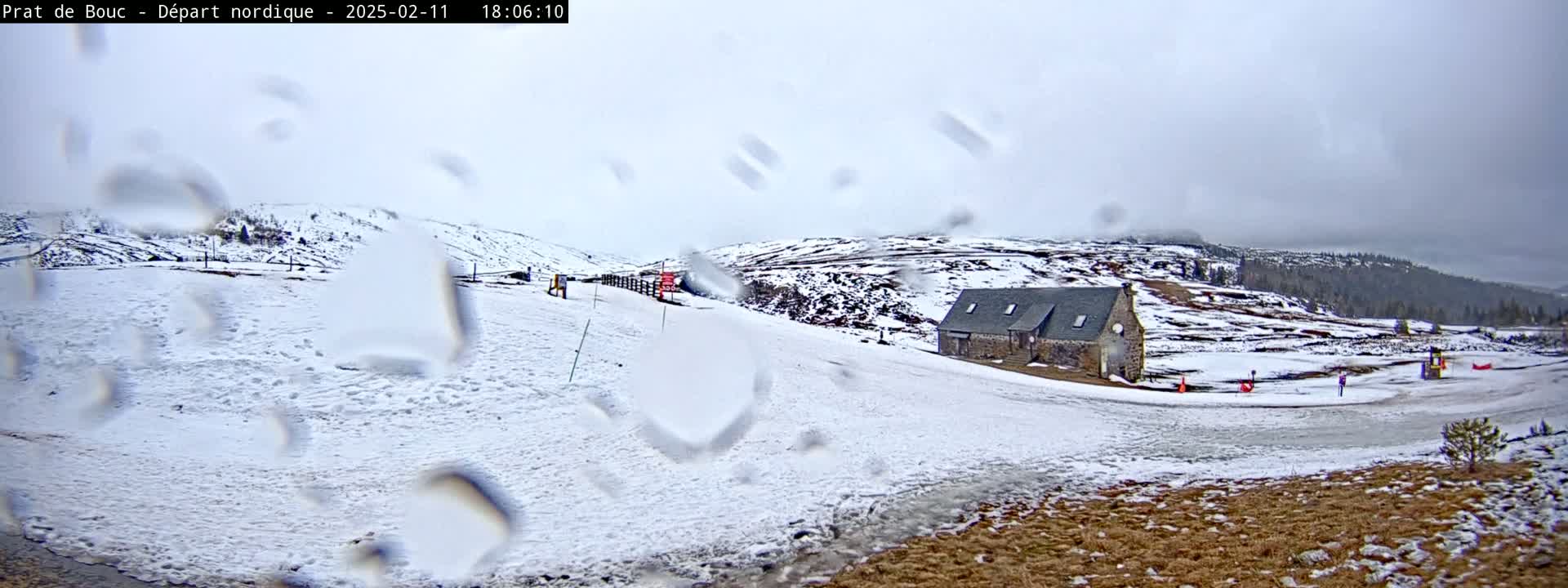A snow-covered landscape with a stone building is seen through rain-covered glass, under an overcast sky.