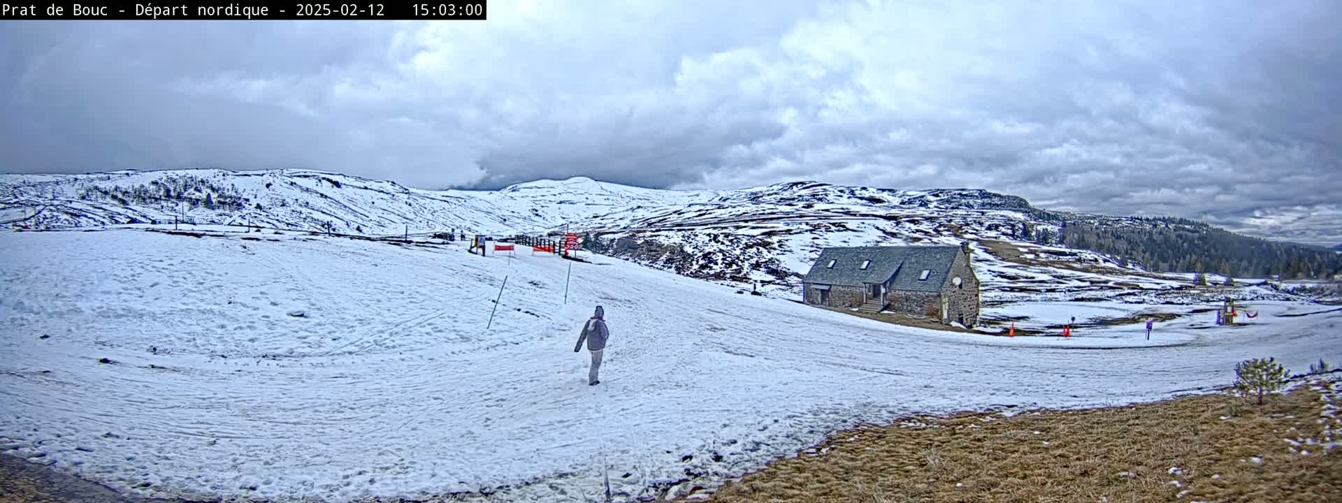 A person walks across a snow-covered hillside towards a stone building under a cloudy sky.