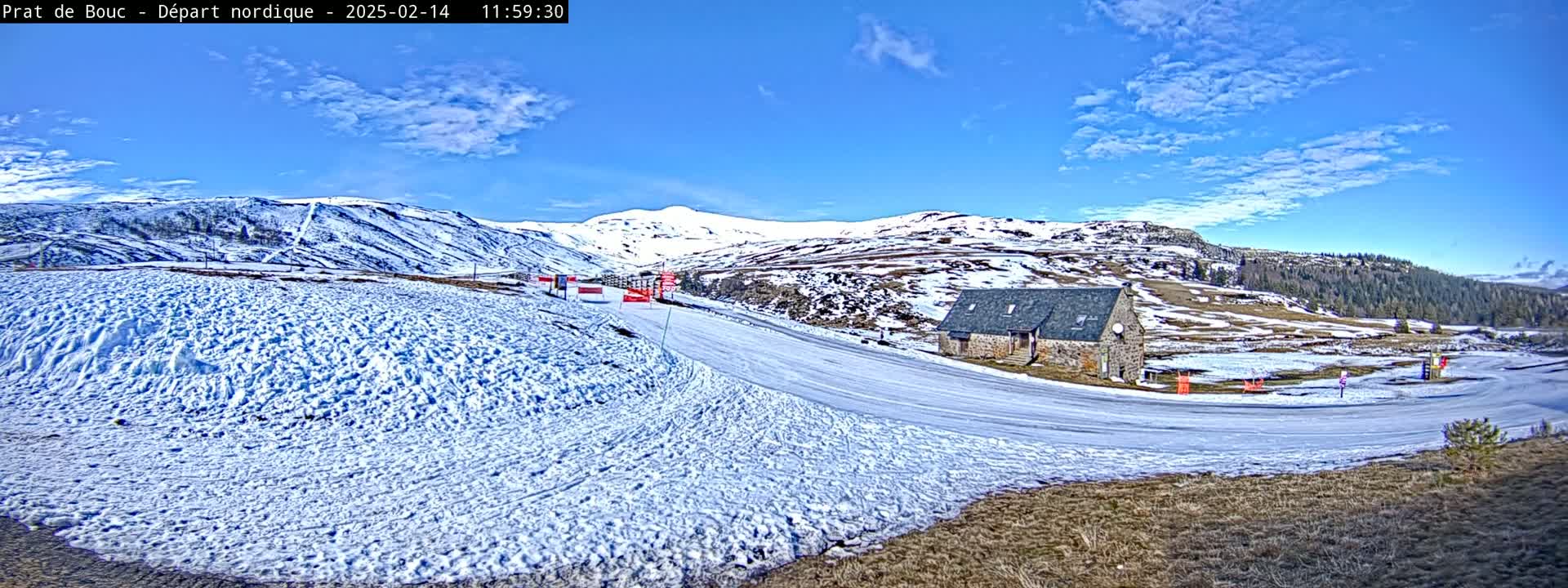 A snow-covered landscape with a stone building near a groomed ski trail under a mostly sunny sky.