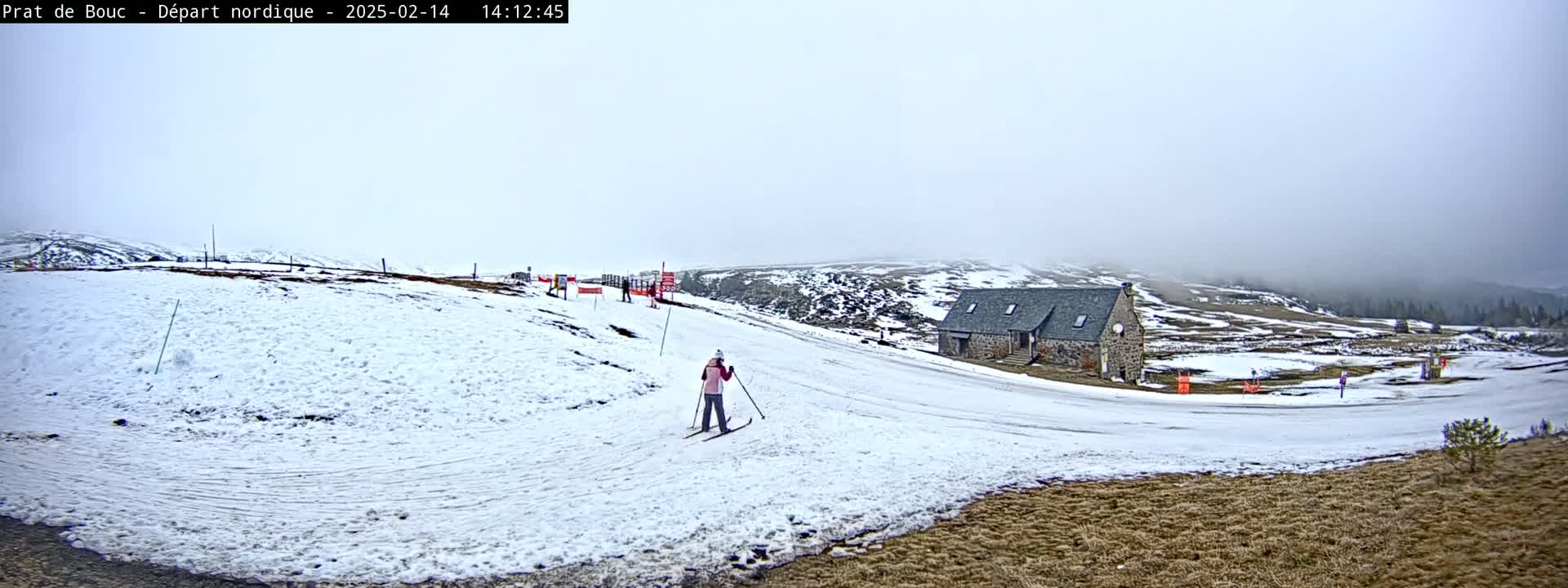 A person cross-country skis on a snow-covered hill near a stone building in foggy conditions.