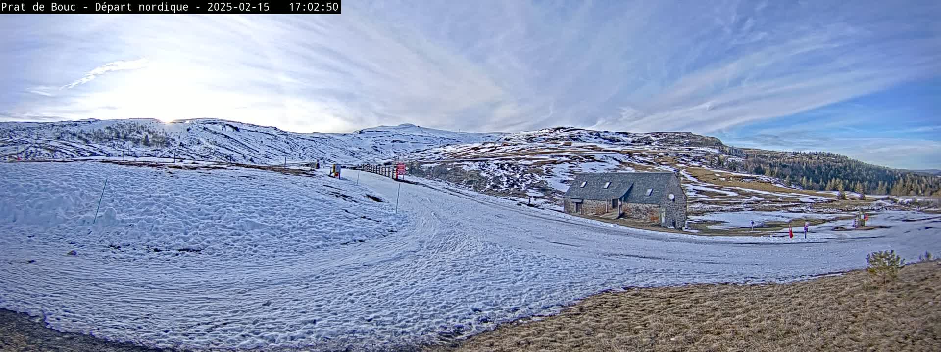 A snow-covered landscape with a small stone building sits below a partly sunny sky, featuring rolling hills and some sparse trees in the distance.