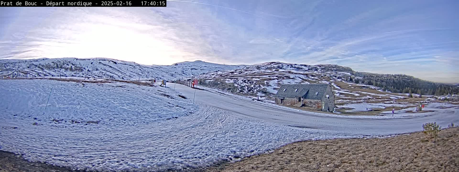 A snow-covered landscape with a small stone building near a road curving through a valley under a partly cloudy, bright sky.