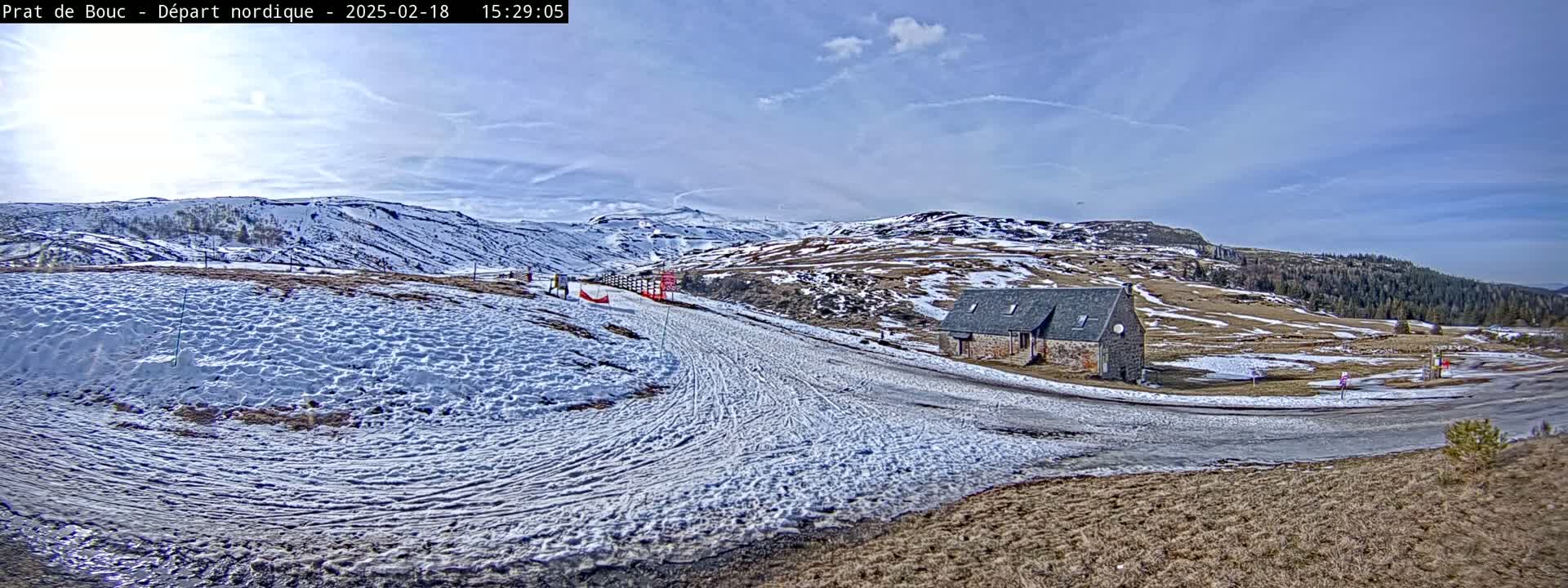 A snow-covered landscape with a small stone building, a groomed ski trail, and distant mountains under a mostly sunny sky.