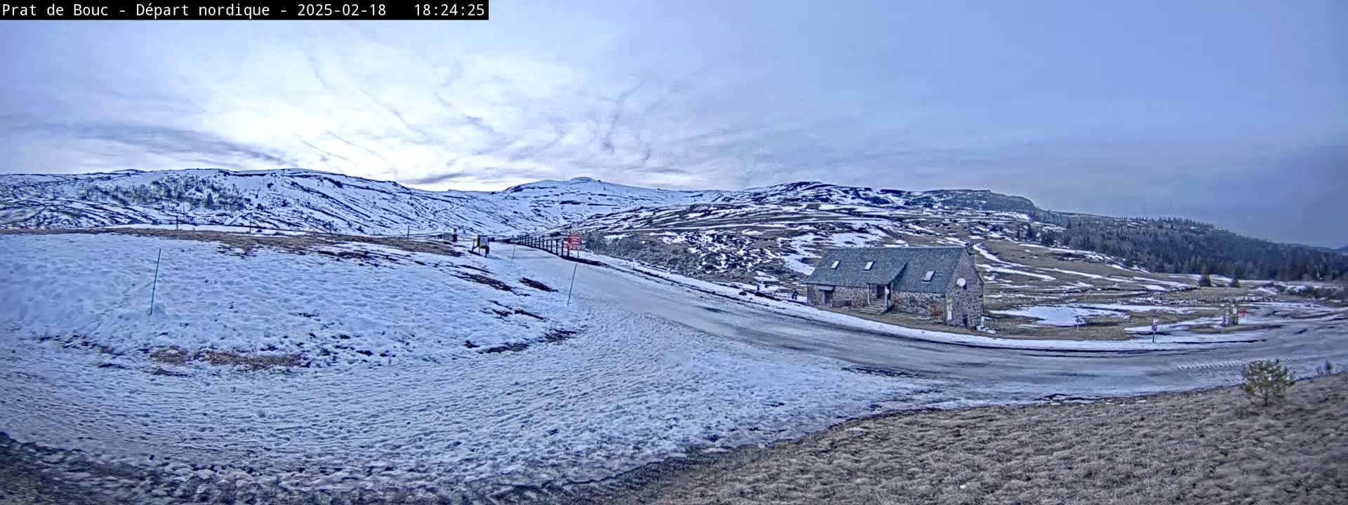A snow-covered landscape with a small stone building near a winding road, under a partly cloudy sky.