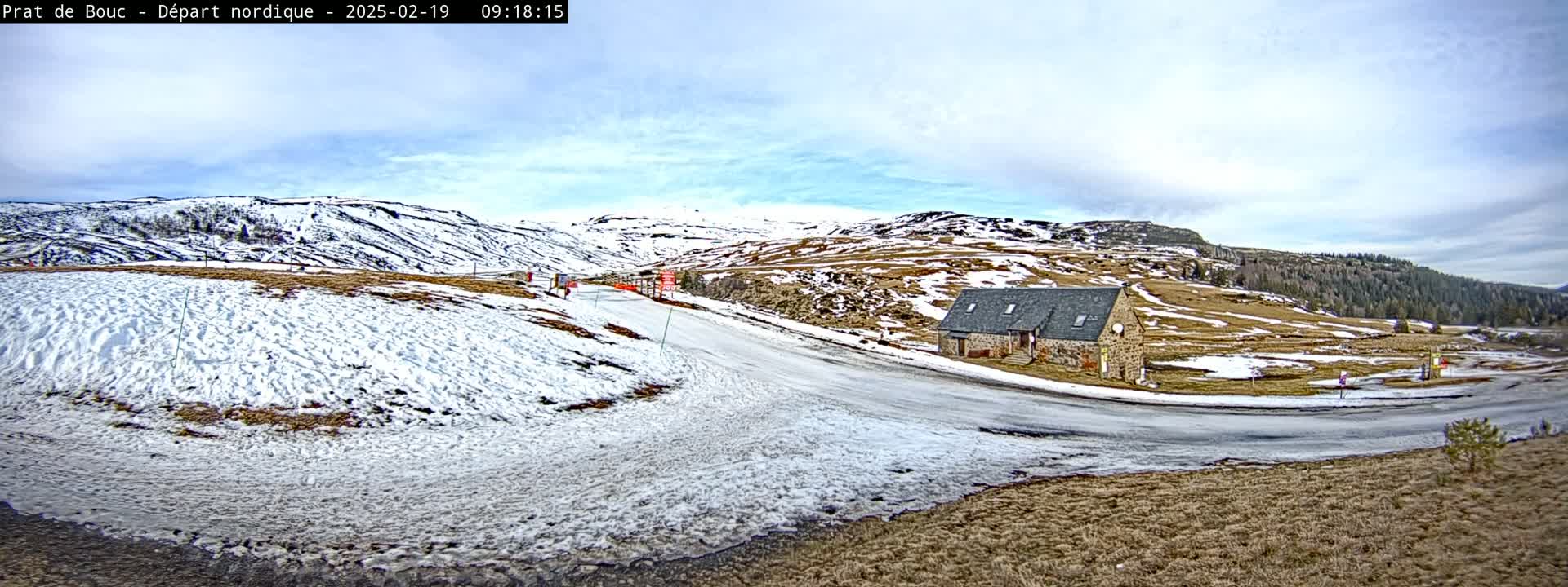 A snow-covered landscape with a stone building near a road curves around a hillside under a partly cloudy sky.