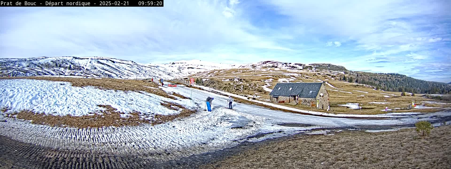 A partly cloudy day reveals a snow-dusted landscape with a stone building near a roadway, where a few people are visible.