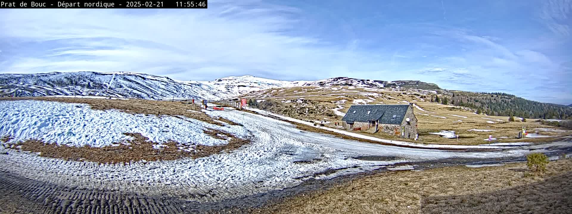A partly sunny day reveals a snow-dusted landscape with a stone building near a road winding through hills, some of which are covered in snow.