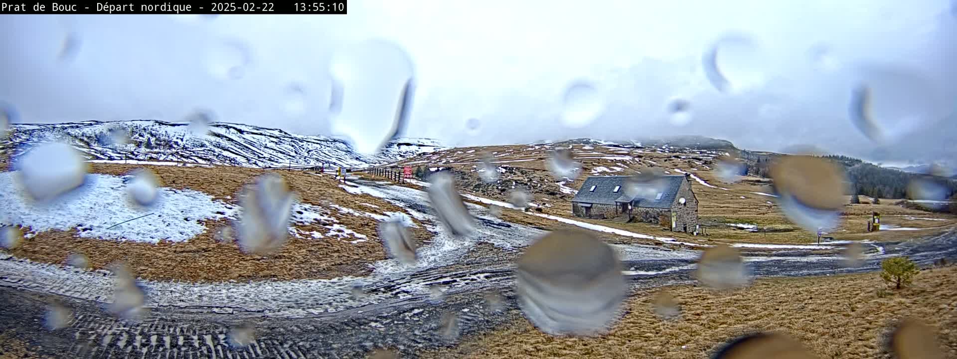 A stone building sits on a snow-dusted hillside under a cloudy sky, viewed through rain-covered glass.
