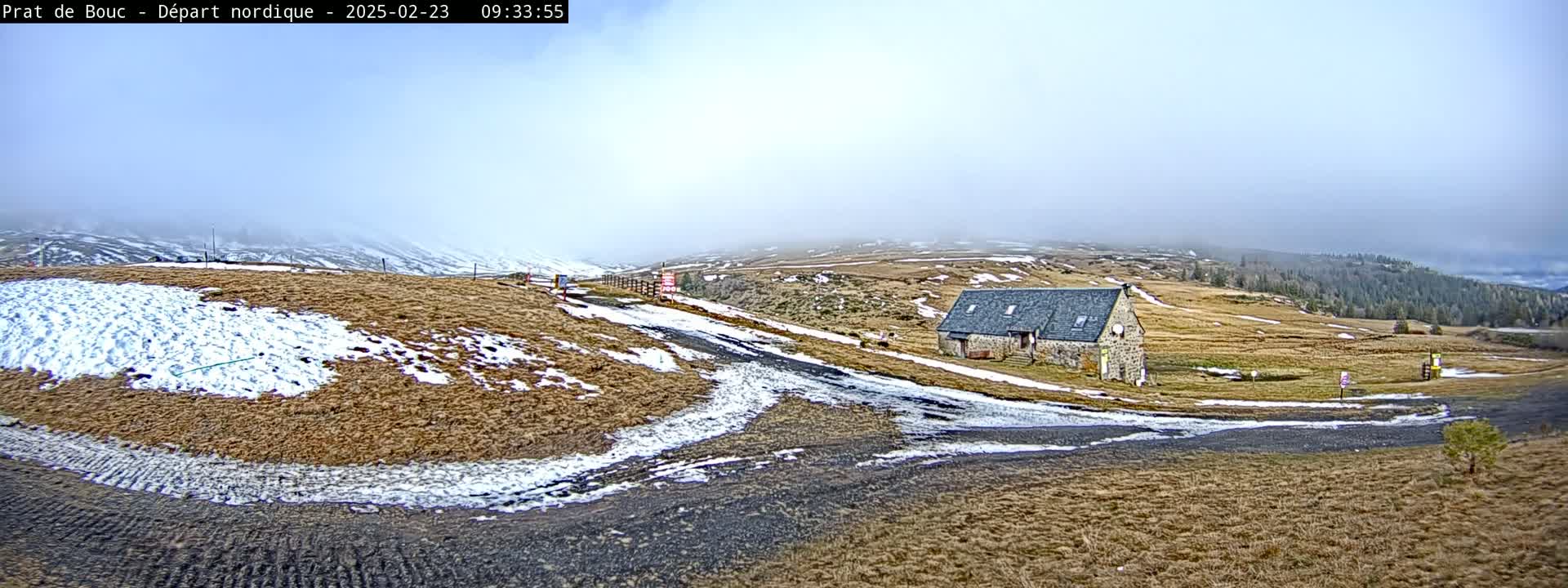 A stone building sits on a sparsely vegetated hillside with patches of snow under a foggy sky.