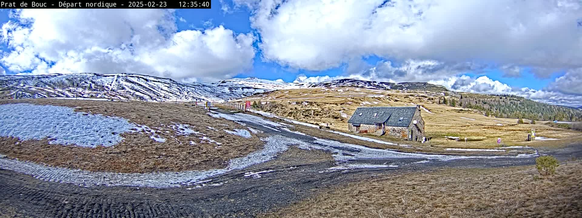 A partly cloudy, bright day reveals a mountain landscape with patches of snow, a small stone building, and a winding dirt road.