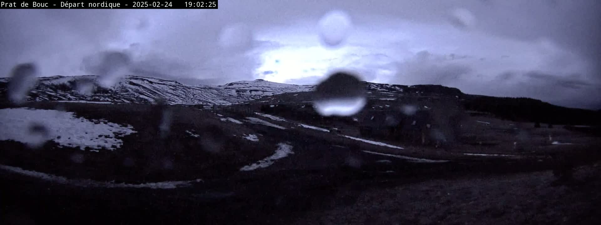 Rain falls on a dark, snow-dusted landscape featuring a hill in the background, viewed through a rain-covered surface.