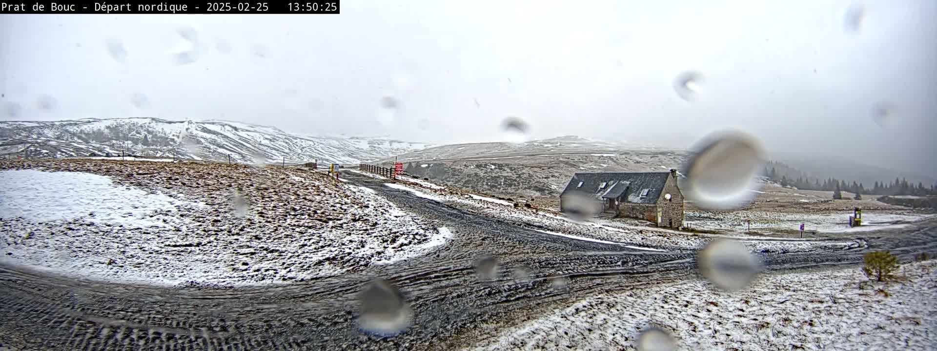 A snow-covered landscape with a small stone building and a dirt road is seen through rain-covered glass in a light snowstorm.
